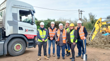 A group of 6 people wearing hard hats and hi-viz jackets are standing on a building site, with a lorry and a digger in the background. One person is holding a spade.