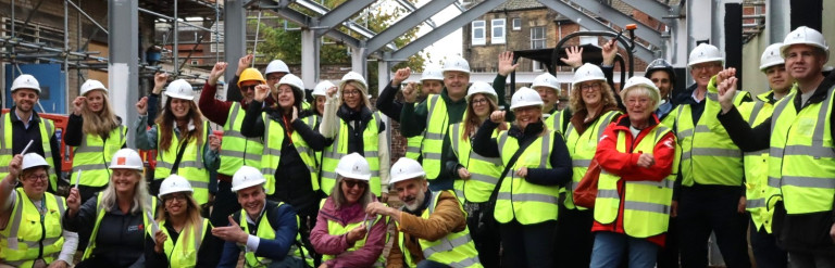 Group photo of people in hi-vis jackets on a building site holding up pens