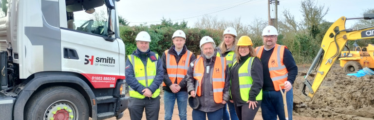 A group of 6 people wearing hard hats and hi-viz jackets are standing on a building site, with a lorry and a digger in the background. One person is holding a spade.